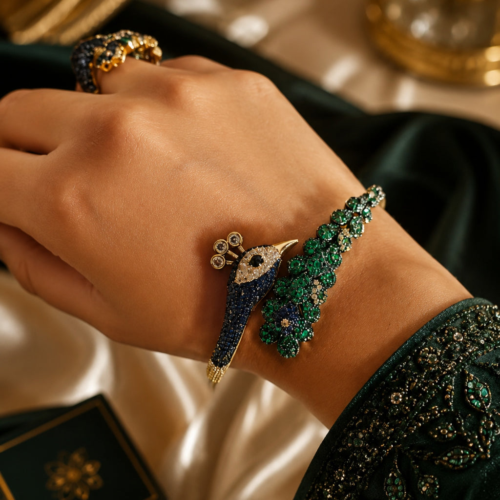 A close-up of a hand wearing a detailed peacock-themed bracelet with blue, green, and gold elements, alongside a matching ring, set against a luxurious dark green and gold fabric background.